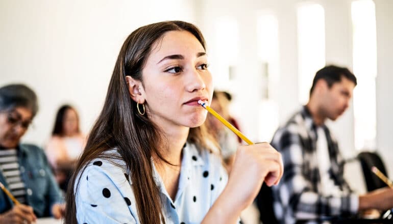 A young college student in class puts a pencil to her lip as she listens intently.