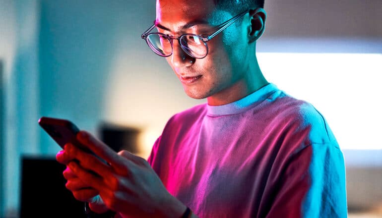A young man looks down at his phone while colorful light shines on him.