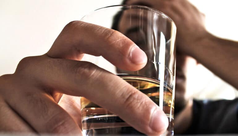 A man holds a glass of liquor through which he can be seen resting his head on his hand.