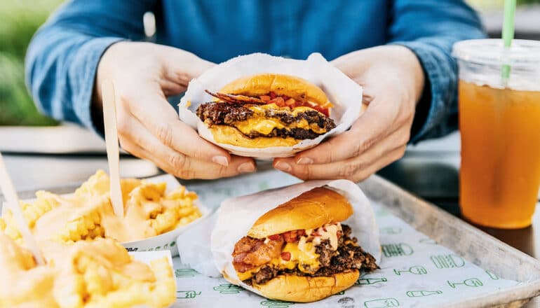 A man eats a cheeseburger while another burger and two orders of fries sit on a tray in front of him.