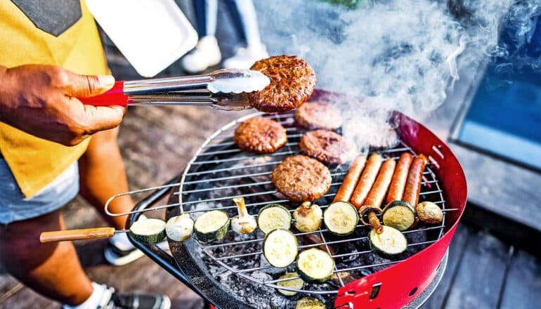 A person grills burgers, hot dogs, and vegetable skewers while outside.