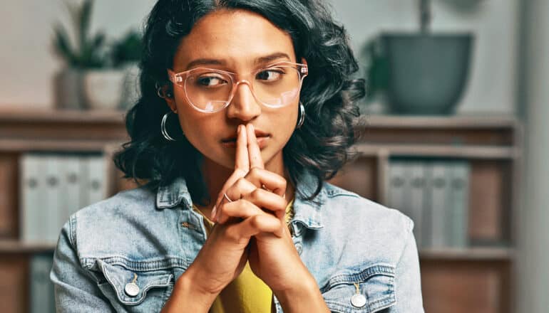 A young woman sitting at a desk presses her hands together and raises them to her lips, looking anxious.