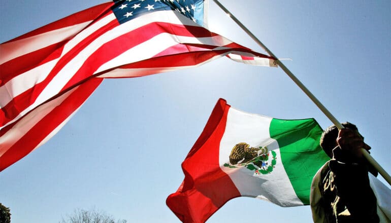 A man holds up the United States flag and the Mexican flag against a blue sky.