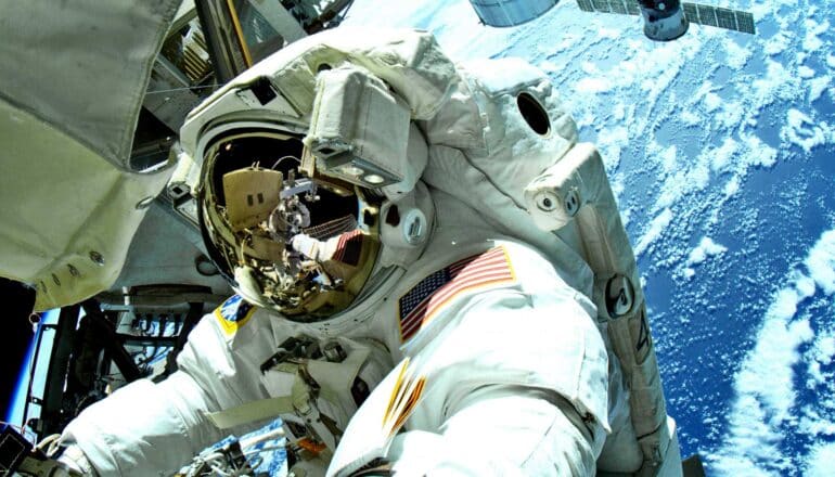 An astronaut works in space near the ISS wearing a NASA spacesuit with the Earth visible in the background.