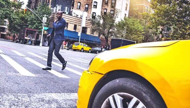 A man walks in front of a yellow taxi while crossing the street.