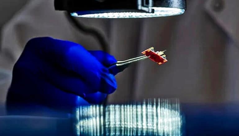 A researcher wearing blue gloves holds the small red sensor under a light using tweezers.