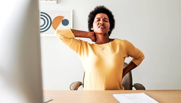 A woman sitting in an office chair winces in pain and holds her neck and lower back.