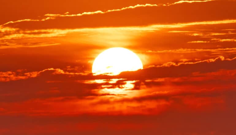 A bright white sun sets behind clouds that are illuminated with red light.