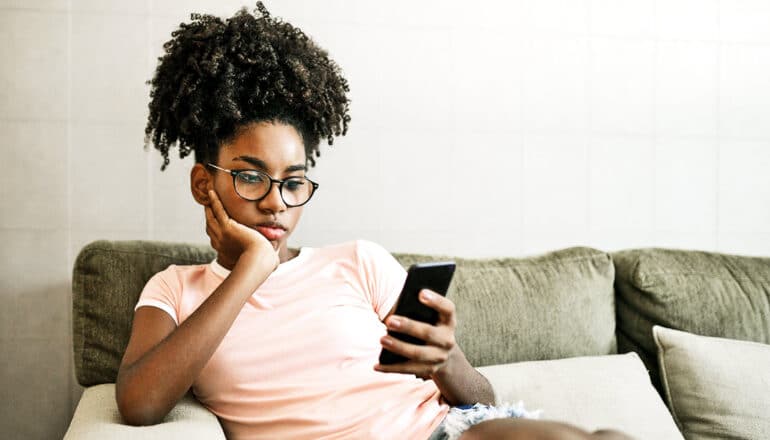A teen girl looks bored while looking at her phone and sitting on a couch.