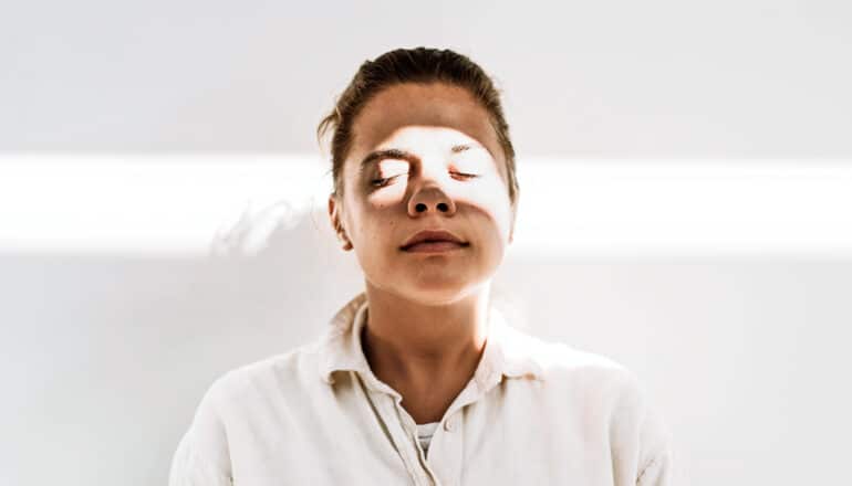 A woman closes her eyes in meditation while a beam of light shines on her face.