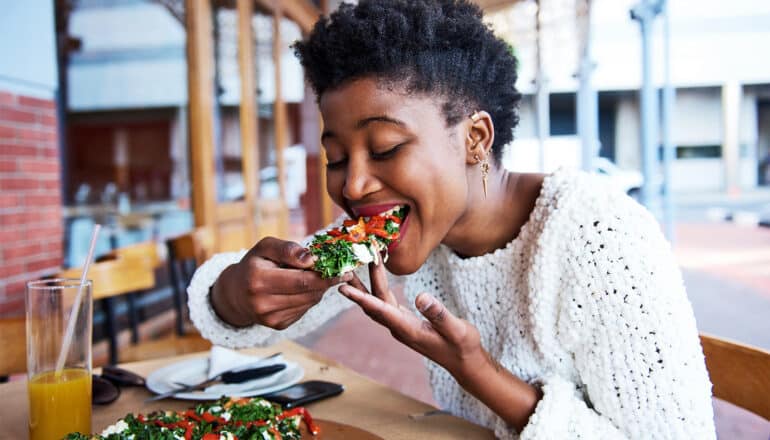 A woman takes a bite of a slice of vegetarian pizza while eating outdoors.