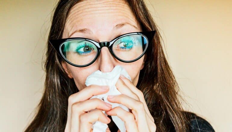 A woman wearing glasses blows her nose with a tissue.