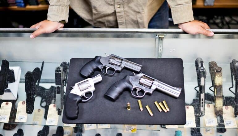 A gun shop worker displays three pistols on a glass case containing more guns.