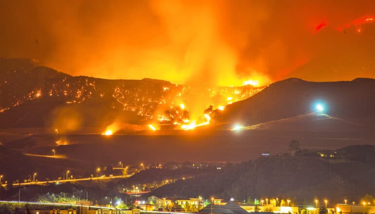 A wildfire burns on the hills behind a small city at night.