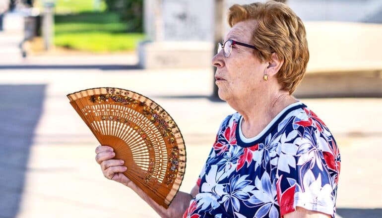 An older woman sits outside in the sun and uses a fan to cool herself.