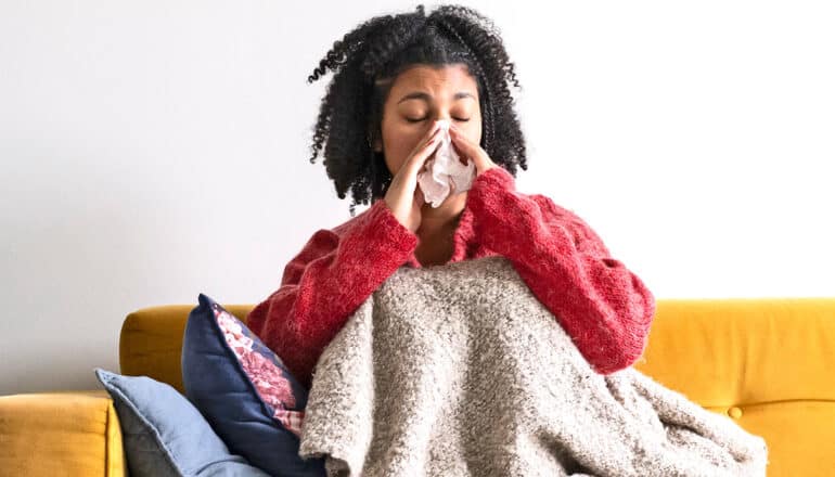 A young woman blows her nose with a tissue while sitting on the couch under a blanket.