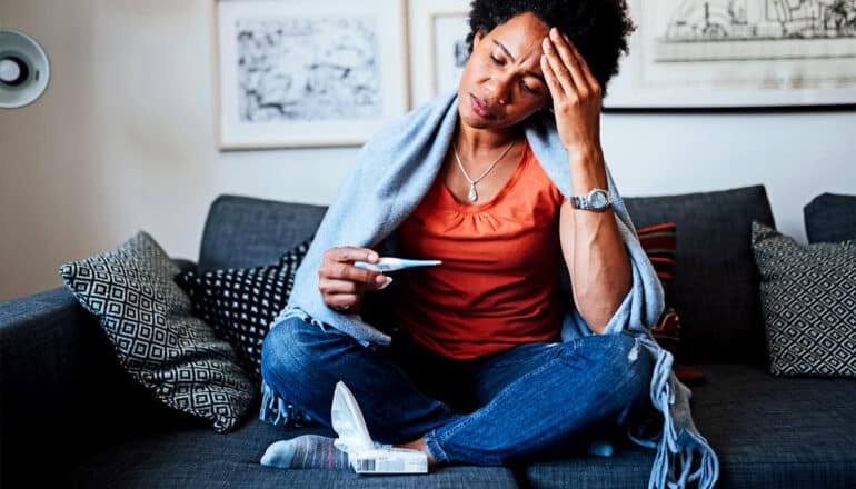 A woman holds her forehead while looking down at a thermometer while sitting under a blanket on a couch and looking uncomfortable.