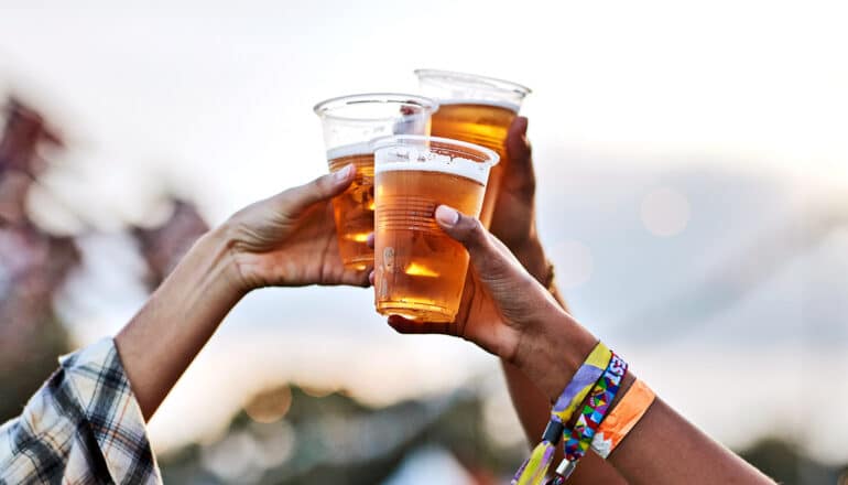 Three people hold up plastic cups of beer in a toast against a white sky.