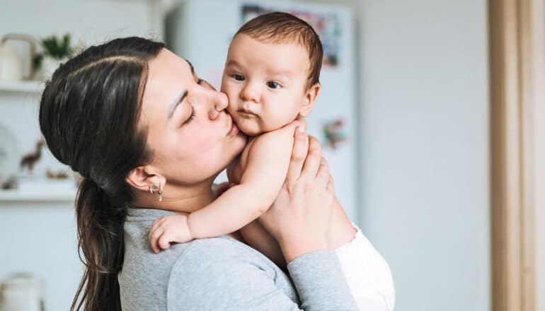 A mother holds up her baby and kisses the baby on the cheek.