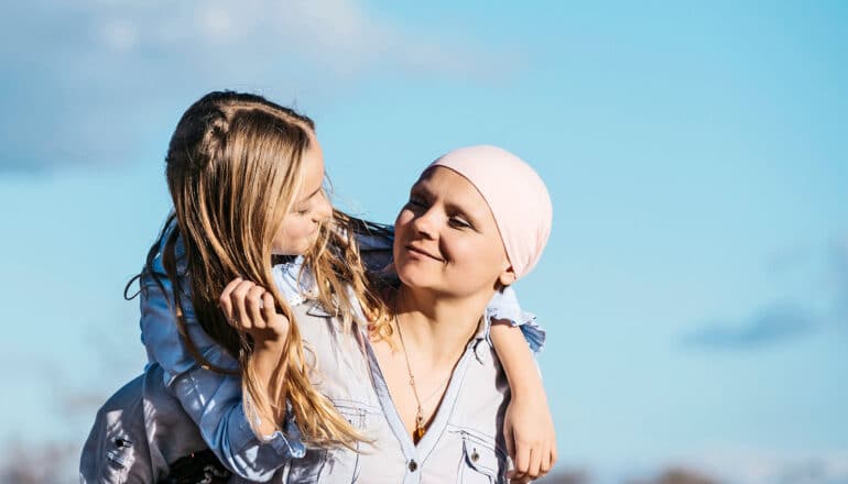 A woman wearing a wrap around her head carries her daughter on her back, who is smiling at her mother.