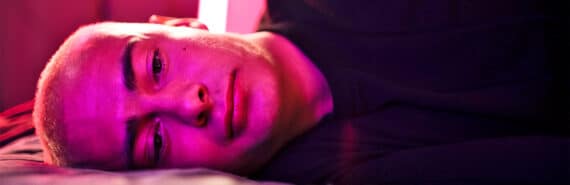 A young man lies on his side in bed and stares into space while illuminated by a pink light.