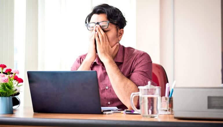 A man sitting at a desk with a laptop on it covers his face with his hands.