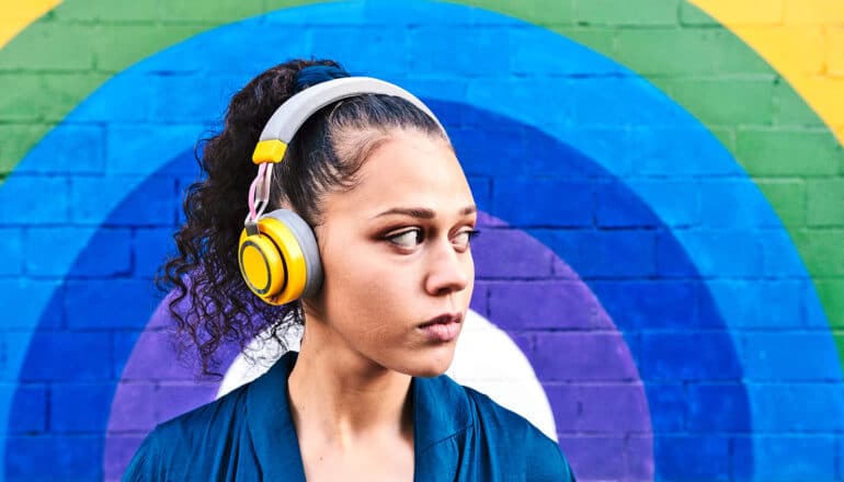 A woman listens to music on large headphones in front of a brick wall painted with colorful arches.