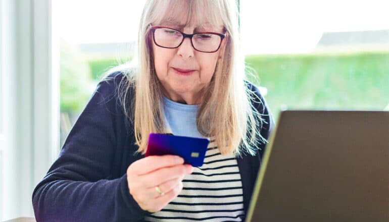 An older woman sits in front of a laptop while looking down at her credit card.