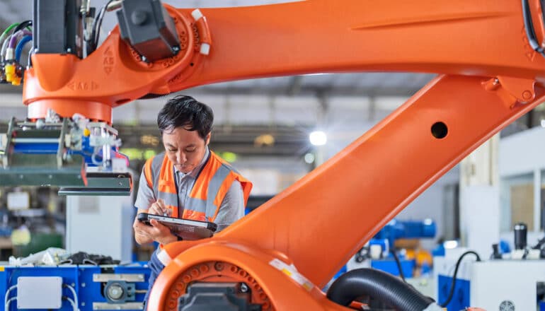 A worker in an orange vest looks down at a tablet computer while standing next to an orange robotic arm in a factory.