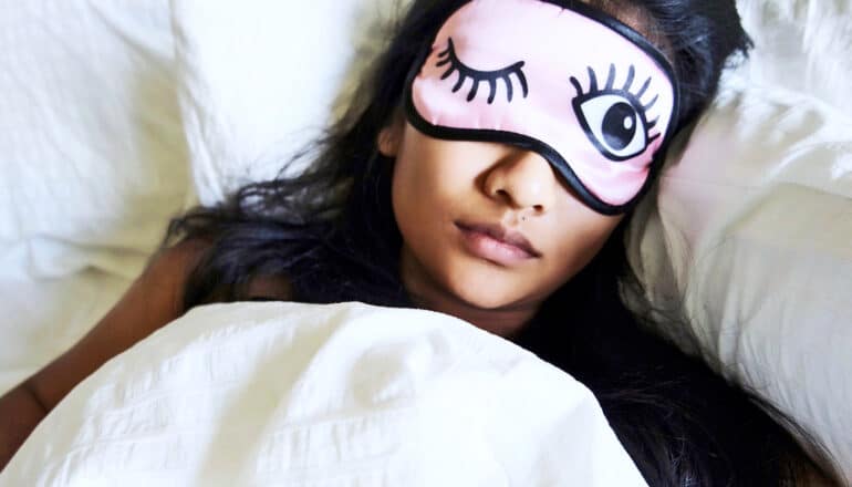 A woman sleeps while wearing a pink sleep mask with one closed and one open eye drawn on it.