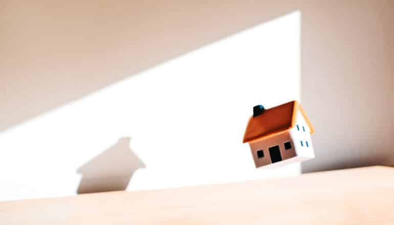 A small toy house floats above a wooden surface and its shadow is cast on the wall behind it.