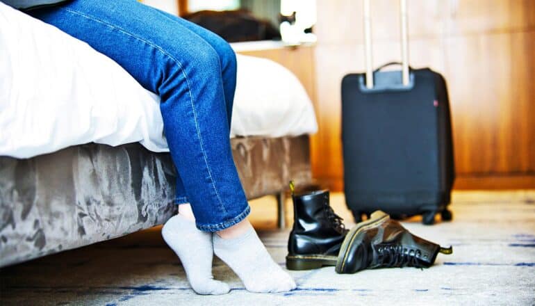 A woman's legs hang off a hotel bed next to her boots and rolling suitcase.