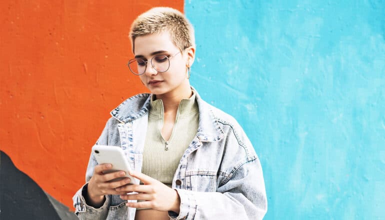 A young woman looks down at her phone while standing in front of a colorful wall.