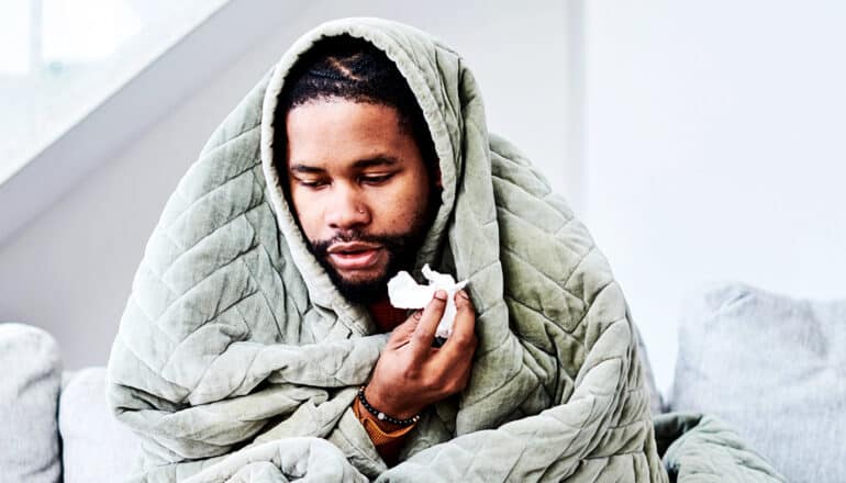 A man sits on a couch under a blanket while holding a tissue in one hand.