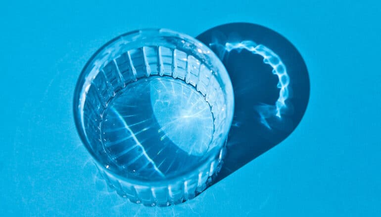 A glass of water sits on a blue surface with light shining through it and casting a shadow.