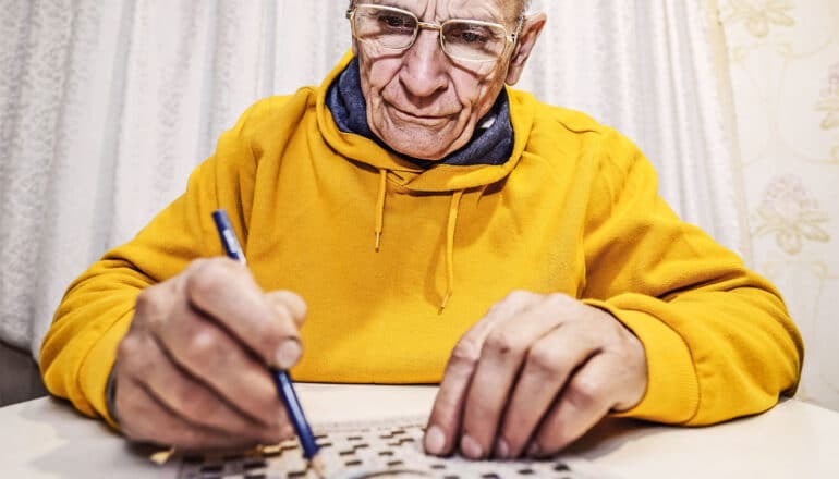 An older man in a yellow hoodie works on a crossword while sitting at a table.