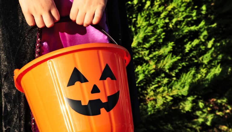 A young girl in a Halloween costume holds an orange plastic bucket with a Jack-o-lantern's face on it.