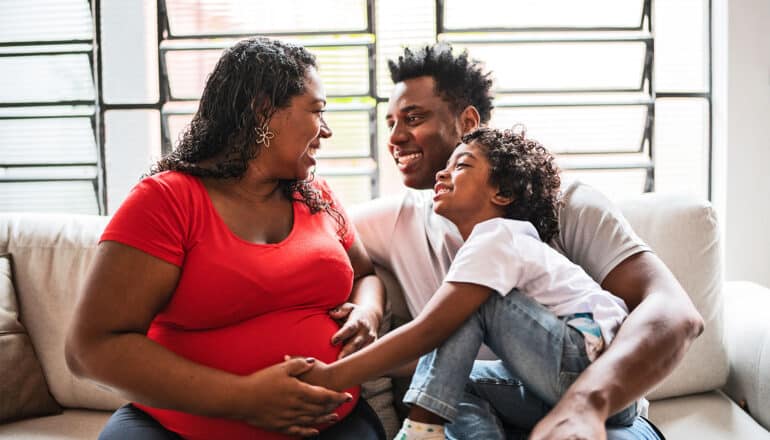 A pregnant woman sits on a couch with her husband and son while they laugh together.