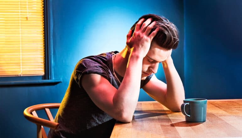 A young man sits at a table with his hands on his head while he stares at a coffee cup.