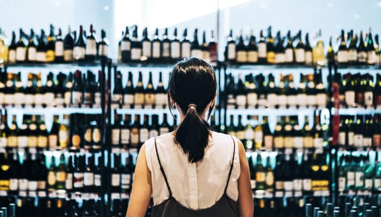 A woman stares at rows and rows of red wine bottles on shelves of a liquor store.