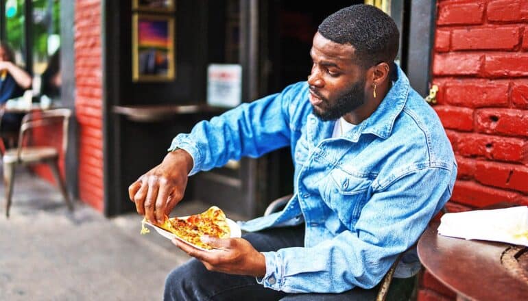 A man sits outside a restaurant with a slice of pizza.