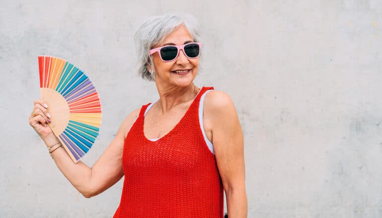 An older woman in sunglasses smiles as she fans herself with a rainbow-colored fan.