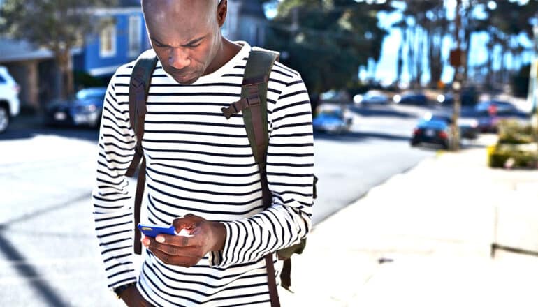 A Black man walking down a street looks down at his phone to order a car from a ride-hailing app.