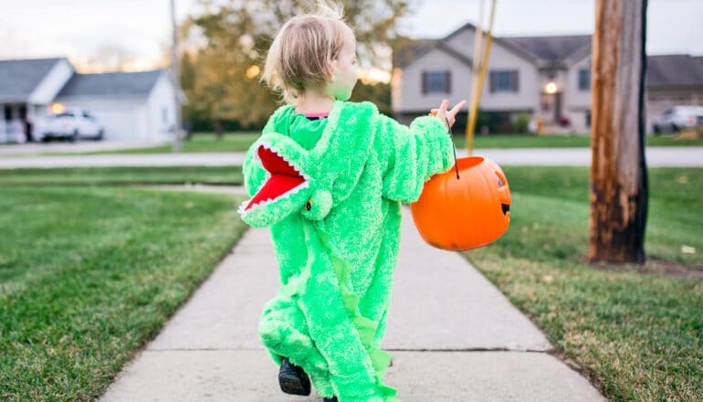 A young child wearing a dinosaur costume walks down a sidewalk while carrying an orange bucket for candy.