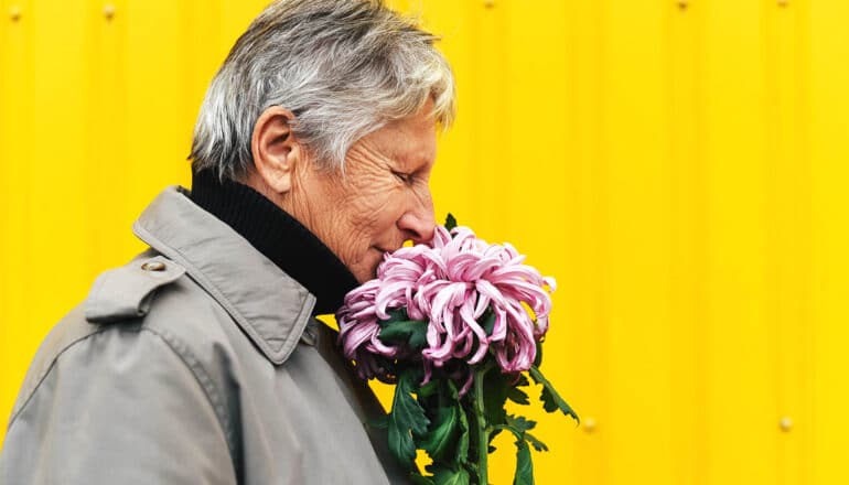 A woman smells flowers while she walks down the street past a yellow wall.