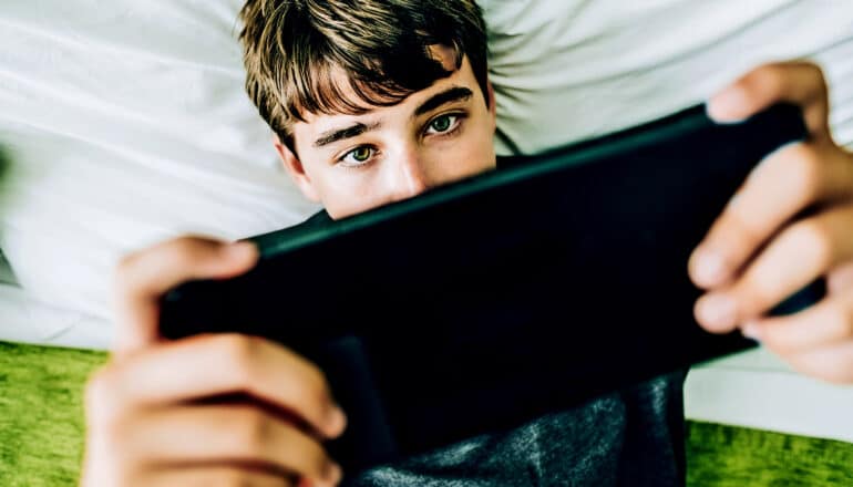 A teen boy holds a game console in his hands while lying down on a bed and playing video games.