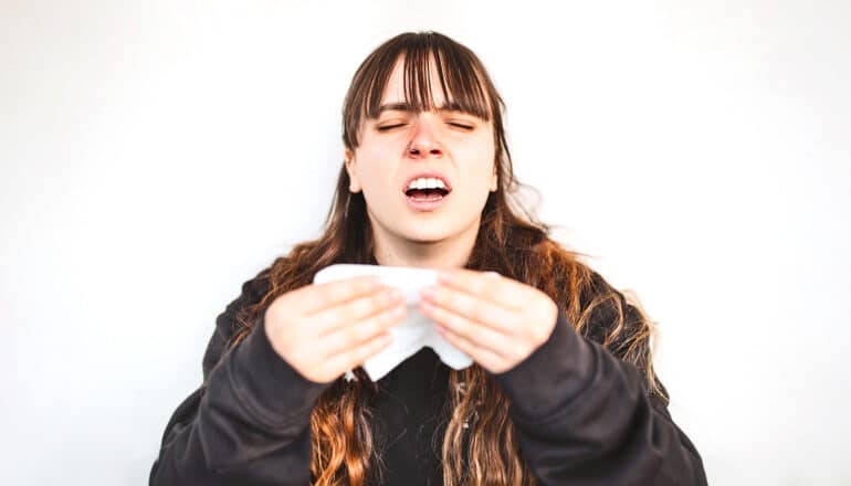 A young woman has her eyes closed and mouth open as she is about to sneeze into a tissue.