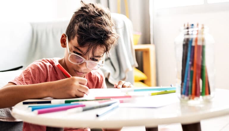 A young boy wearing glasses draws with colored pencils while sitting on the floor at a coffee table.