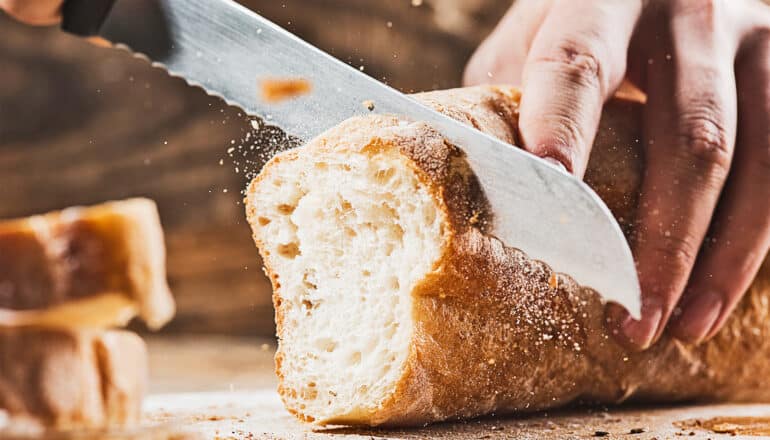 A person uses a knife to cut a loaf of bread as crumbs fly everywhere.