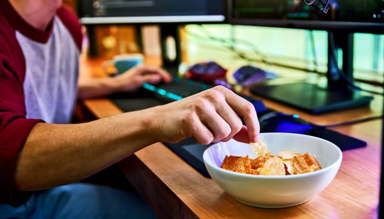 A young man reaches for a potato chip while sitting in front of a computer and gaming.